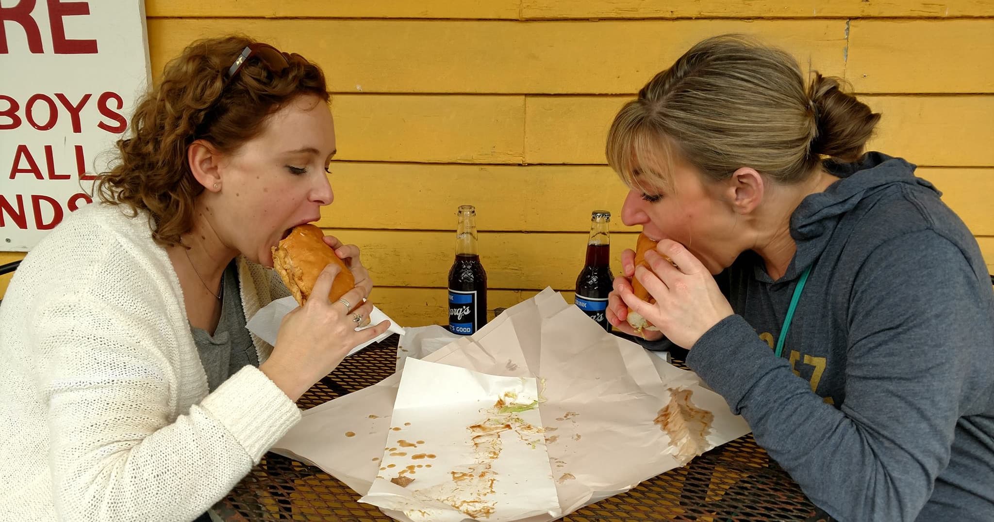 Two women sharing large sandwiches at outdoor market wooden stall