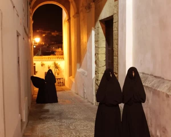Participants in traditional cloaks walking narrow Andalusian street at night