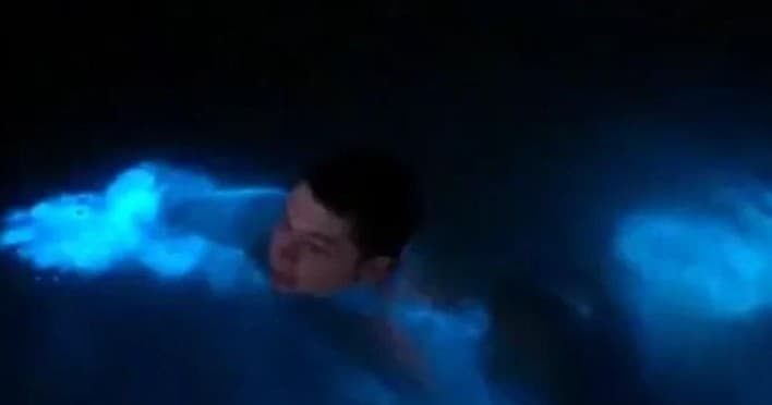 Person swimming in blue bioluminescent surf at night, intimate close-up