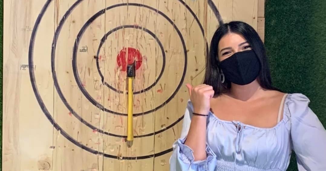 Masked woman posing by bullseye axe throwing target at indoor venue