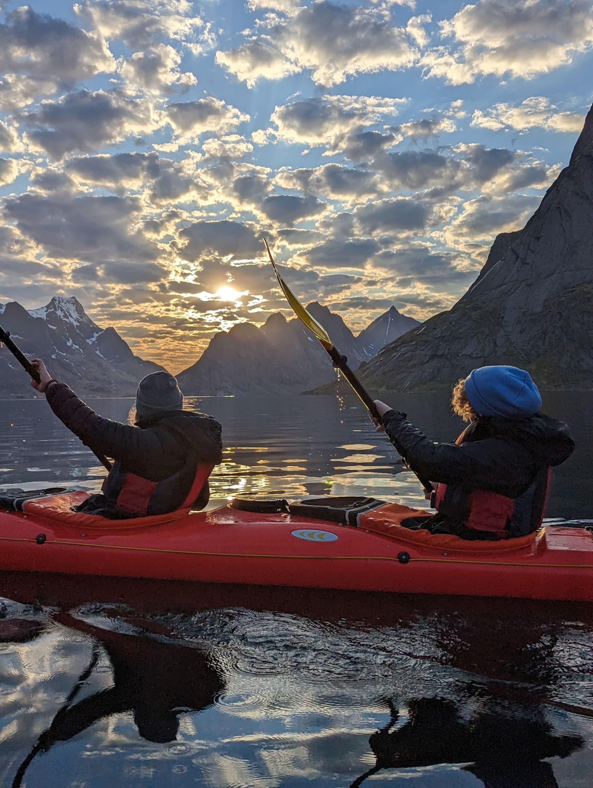Two kayakers paddling at sunrise with mountain reflections in fjord