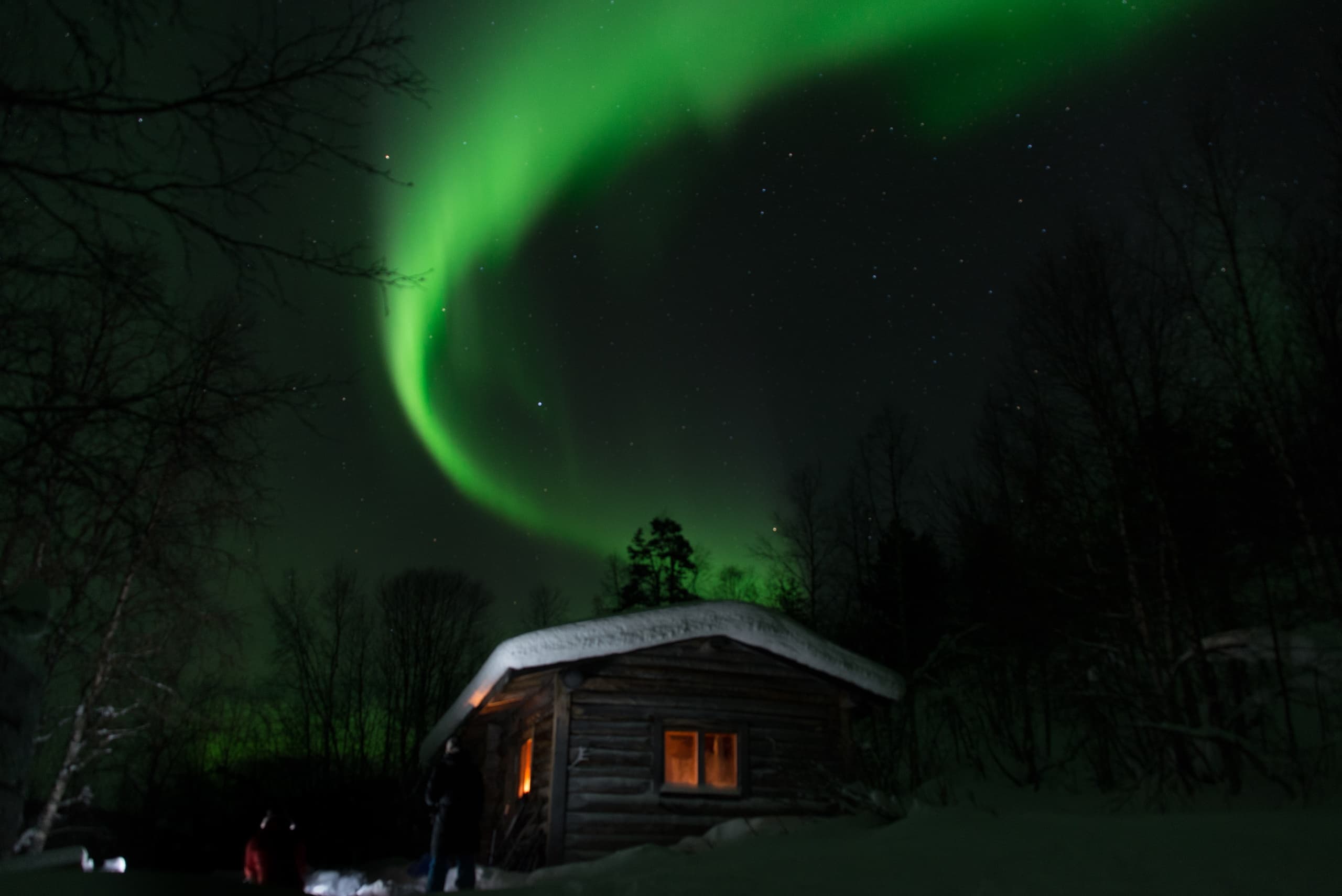 Northern Lights arching over snowy log cabin in Finnish wilderness
