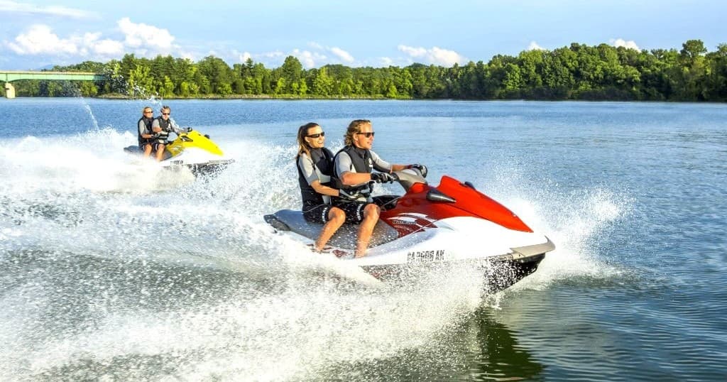 Two people jet skiing on a sunny lake with forested shore