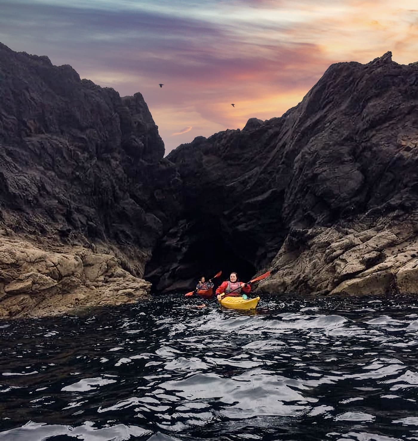 Sea kayakers navigating coastal cliff gorge at sunset