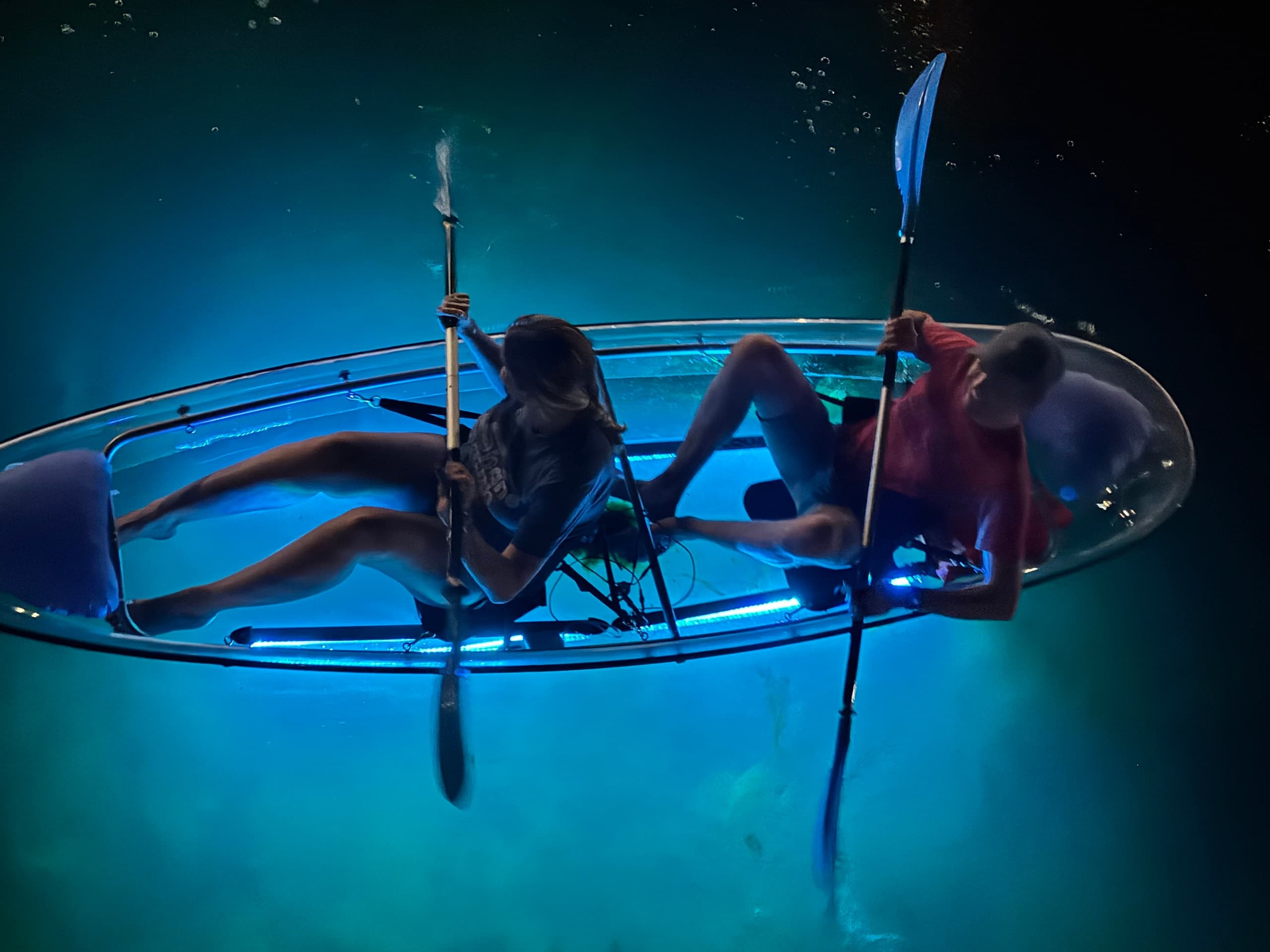Two people kayaking in illuminated clear kayak over turquoise night water