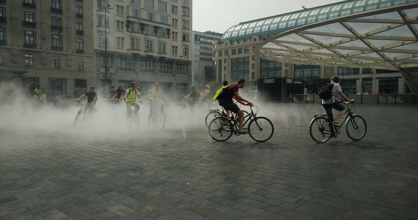 Cyclists riding through misty city plaza near modern glass canopy