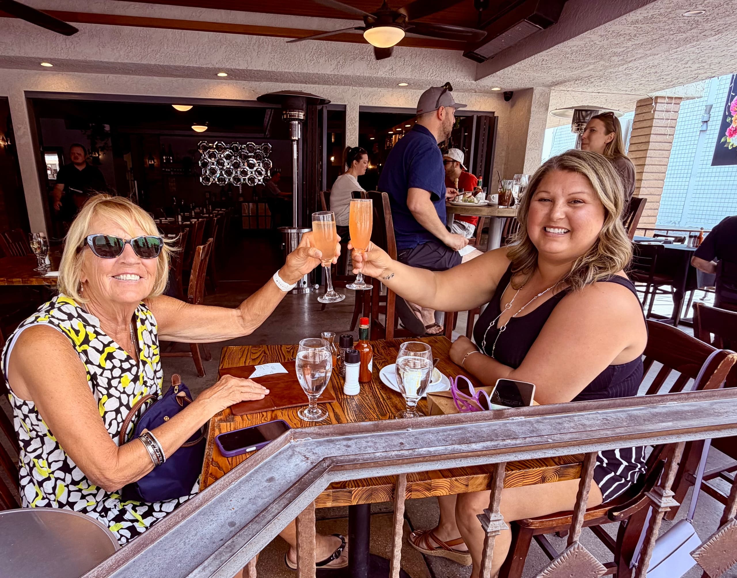 Two women toasting brunch on a sunny restaurant patio outdoors
