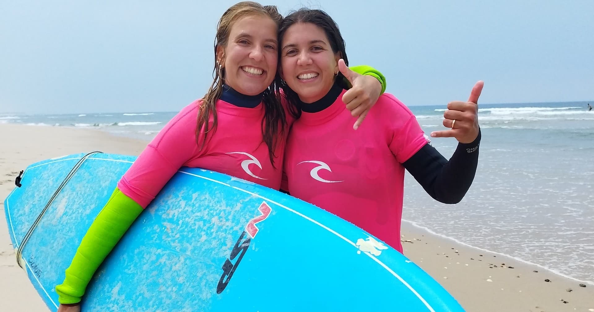 Two surfers in wetsuits posing with surfboard on sandy beach