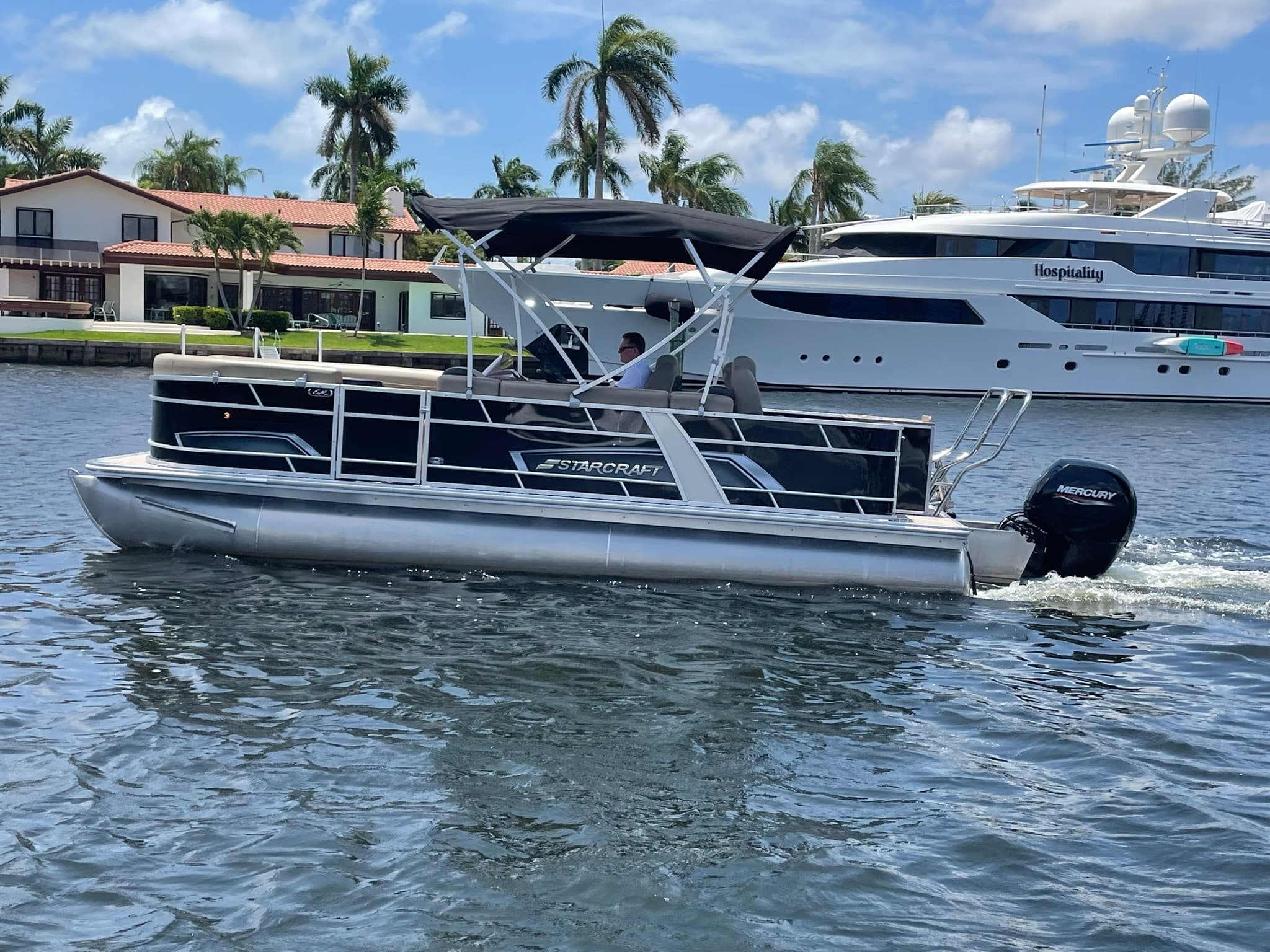 Pontoon boat cruising marina with palm-lined shore and yachts