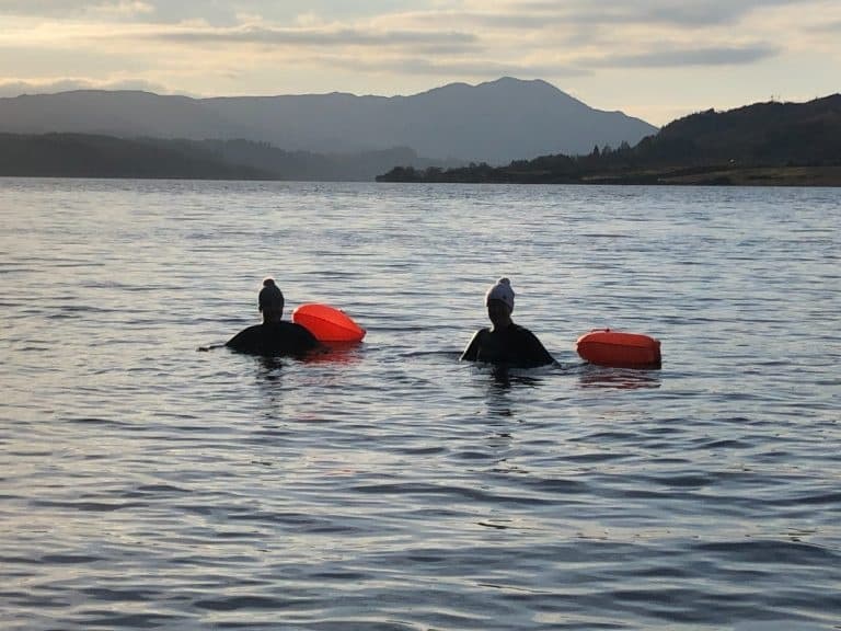 Open-water swimmers with orange buoys on loch at dusk