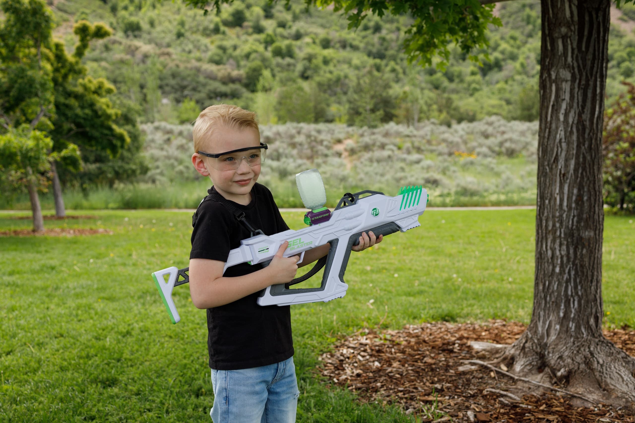 Boy firing gel blaster outdoors in grassy park by a tree