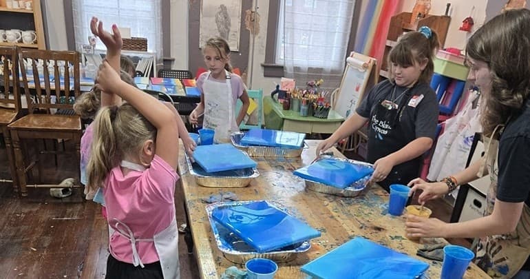 Children painting canvases at long table in indoor art class