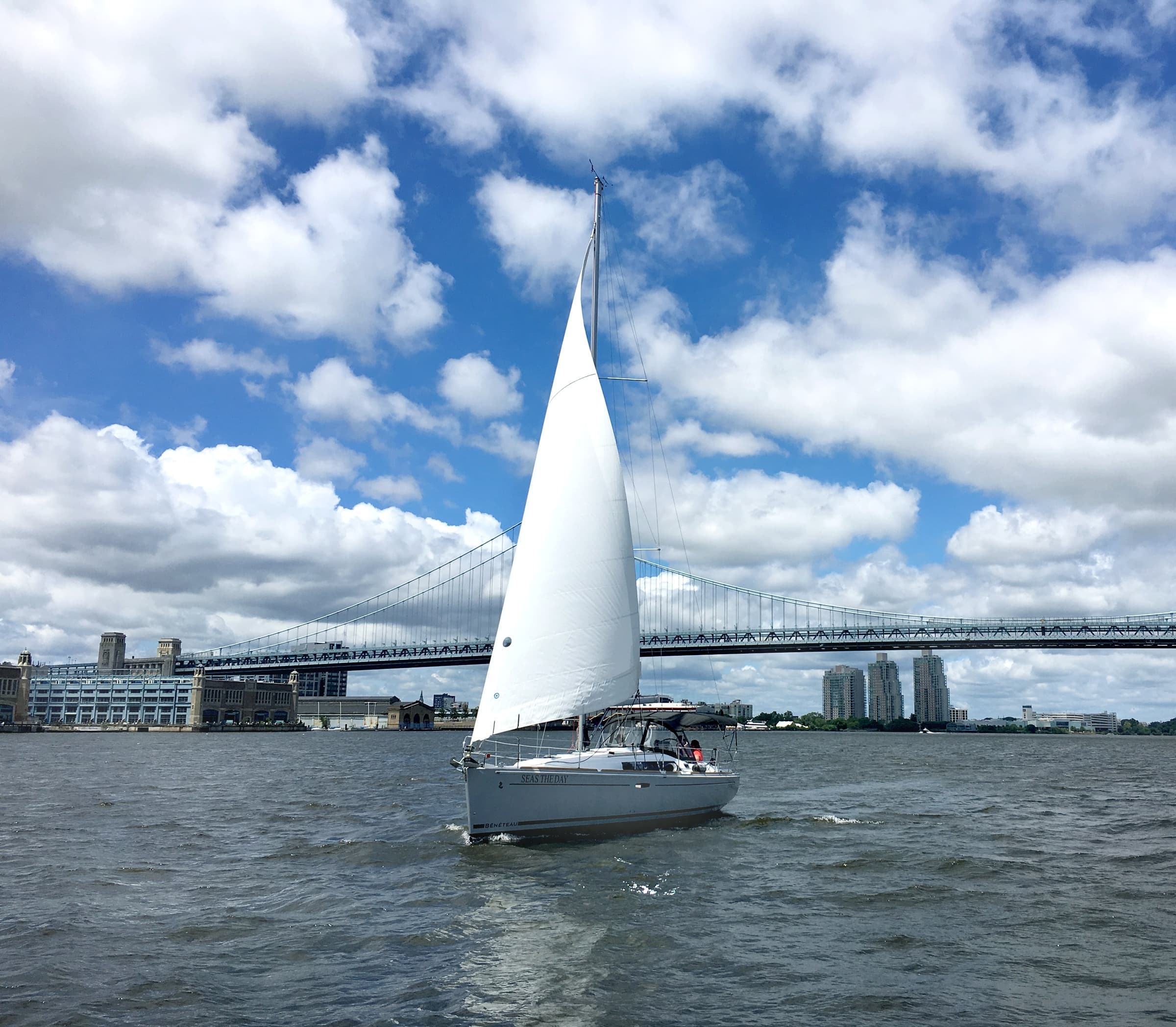 Sailboat under full sail on open water near city bridge