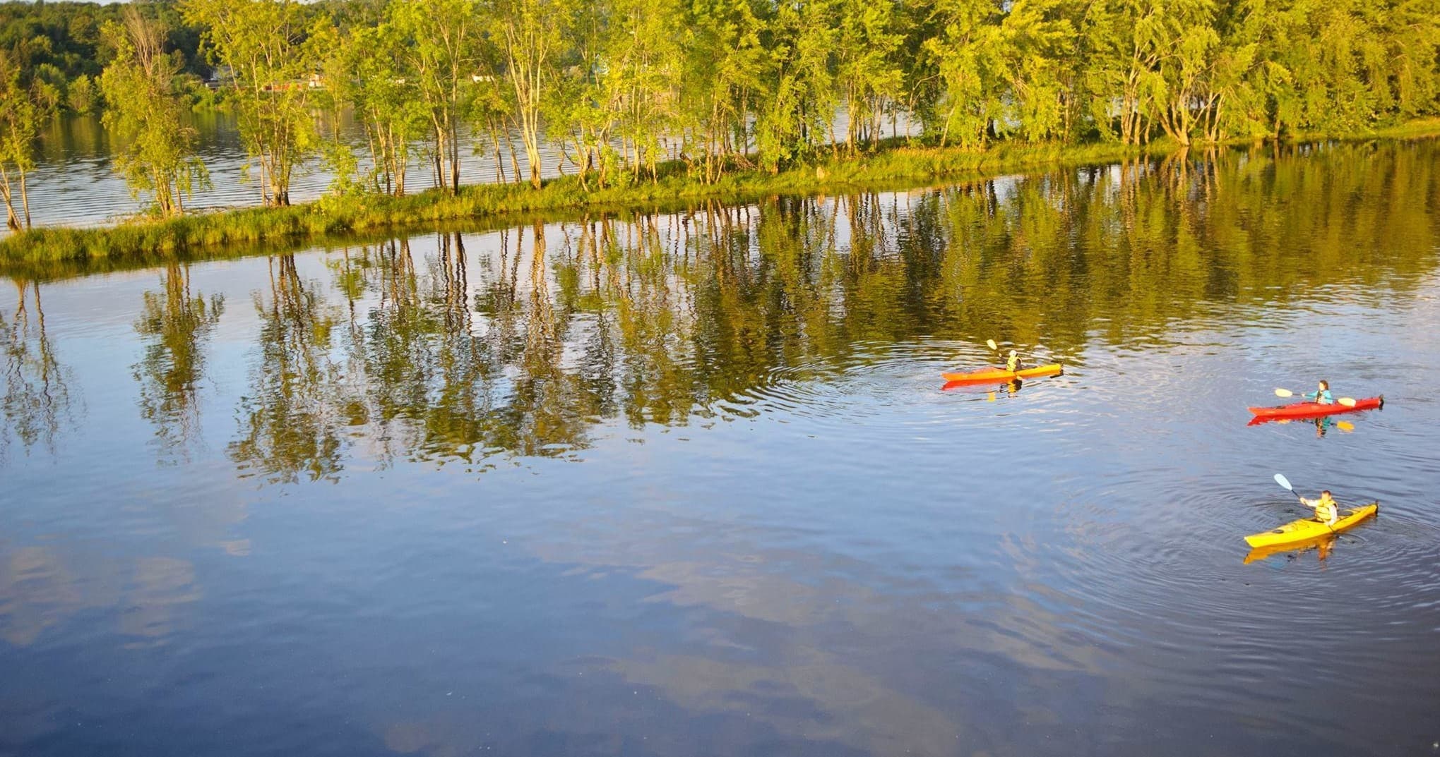 Three kayaks paddling on reflective river by forested shoreline