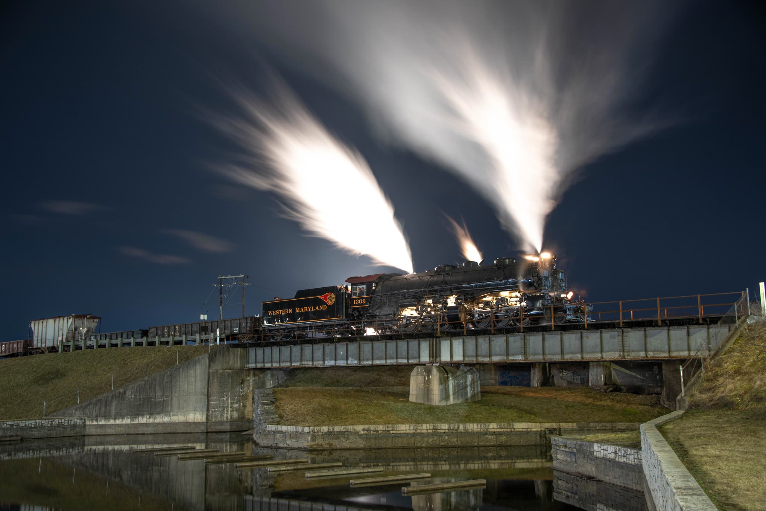 Night steam locomotive with dramatic smoke over riverside bridge