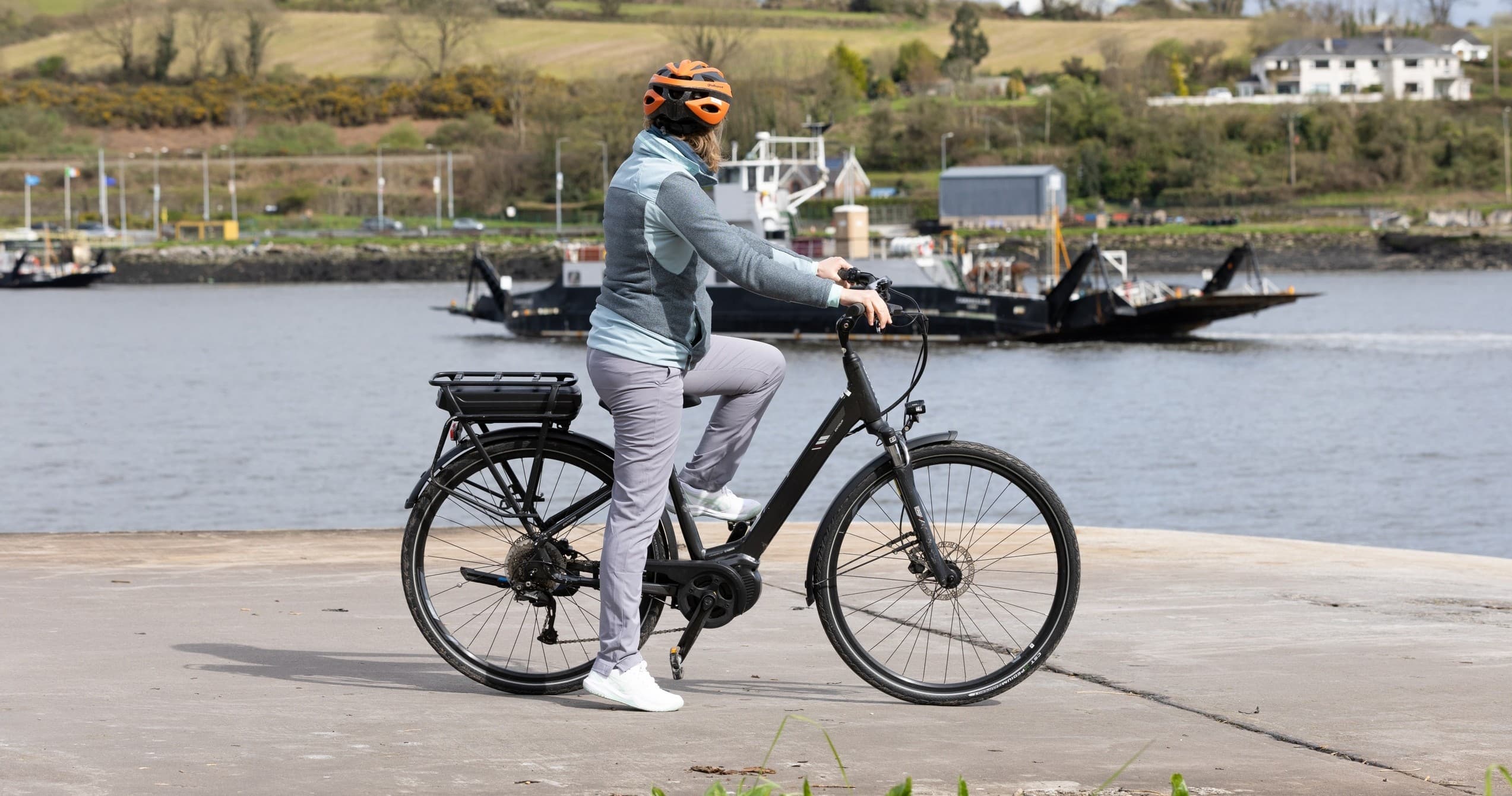 Solo e-bike rider paused by riverside with ferry in background