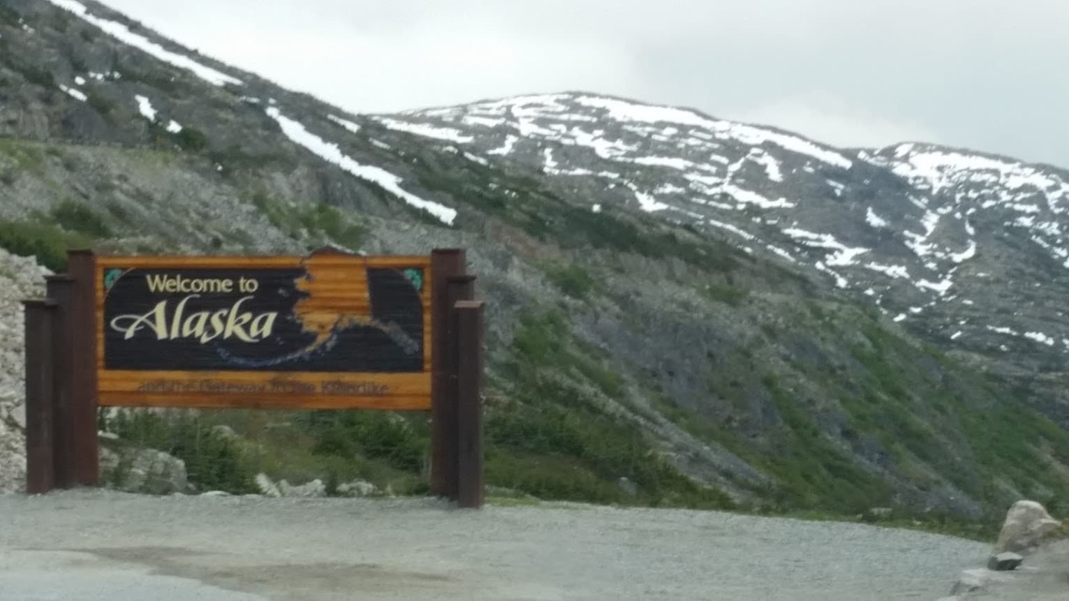 Scenic Alaska welcome sign with snow-capped mountain pass view