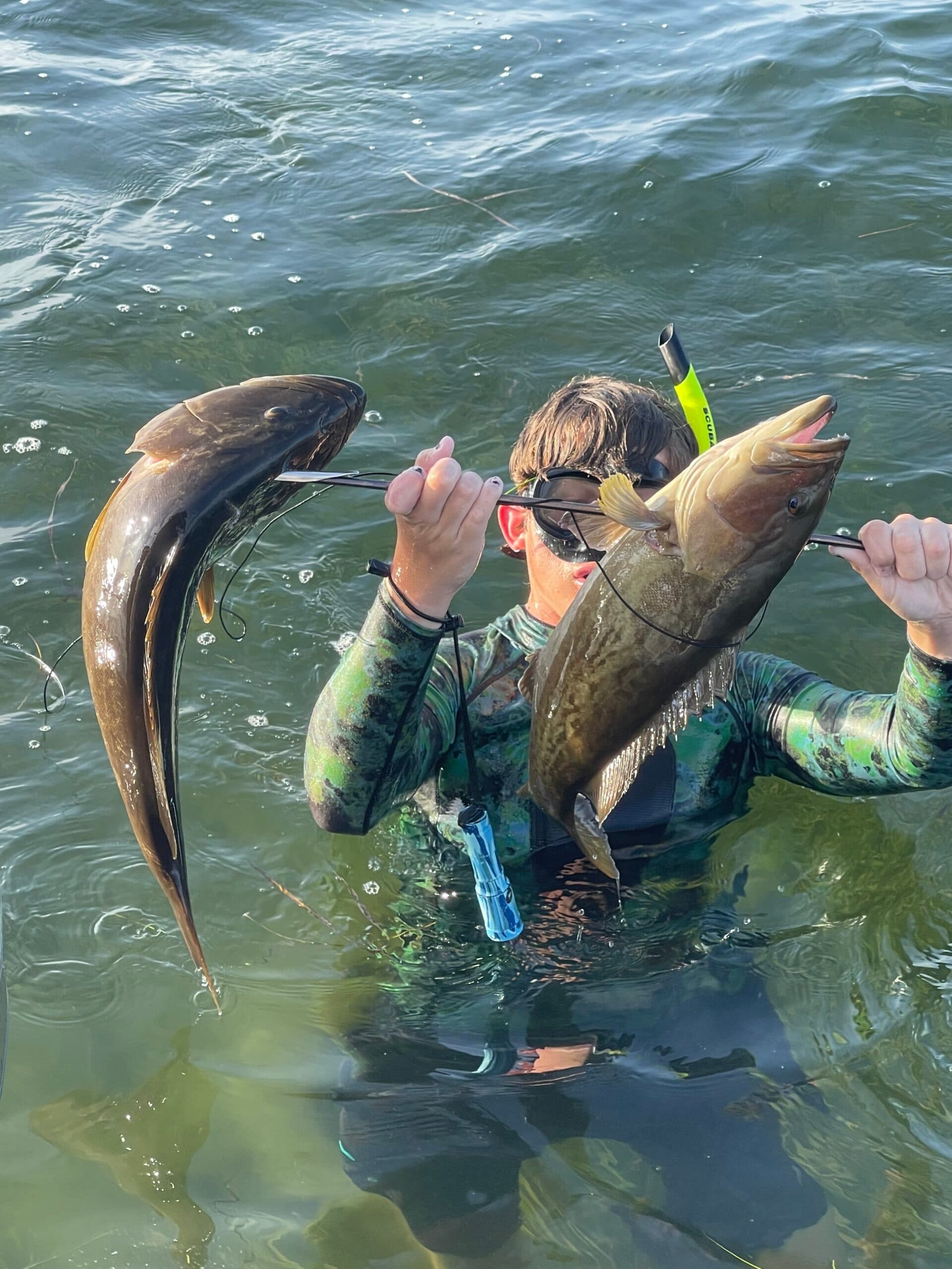 Spearfisher emerging from water holding two fish near reef