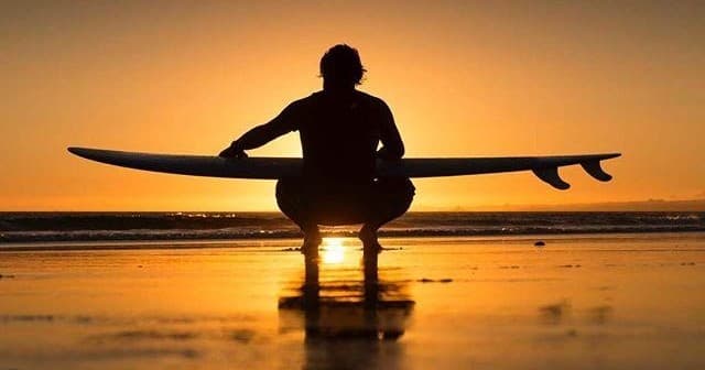 Surfer silhouetted with longboard at sunset on sandy beach