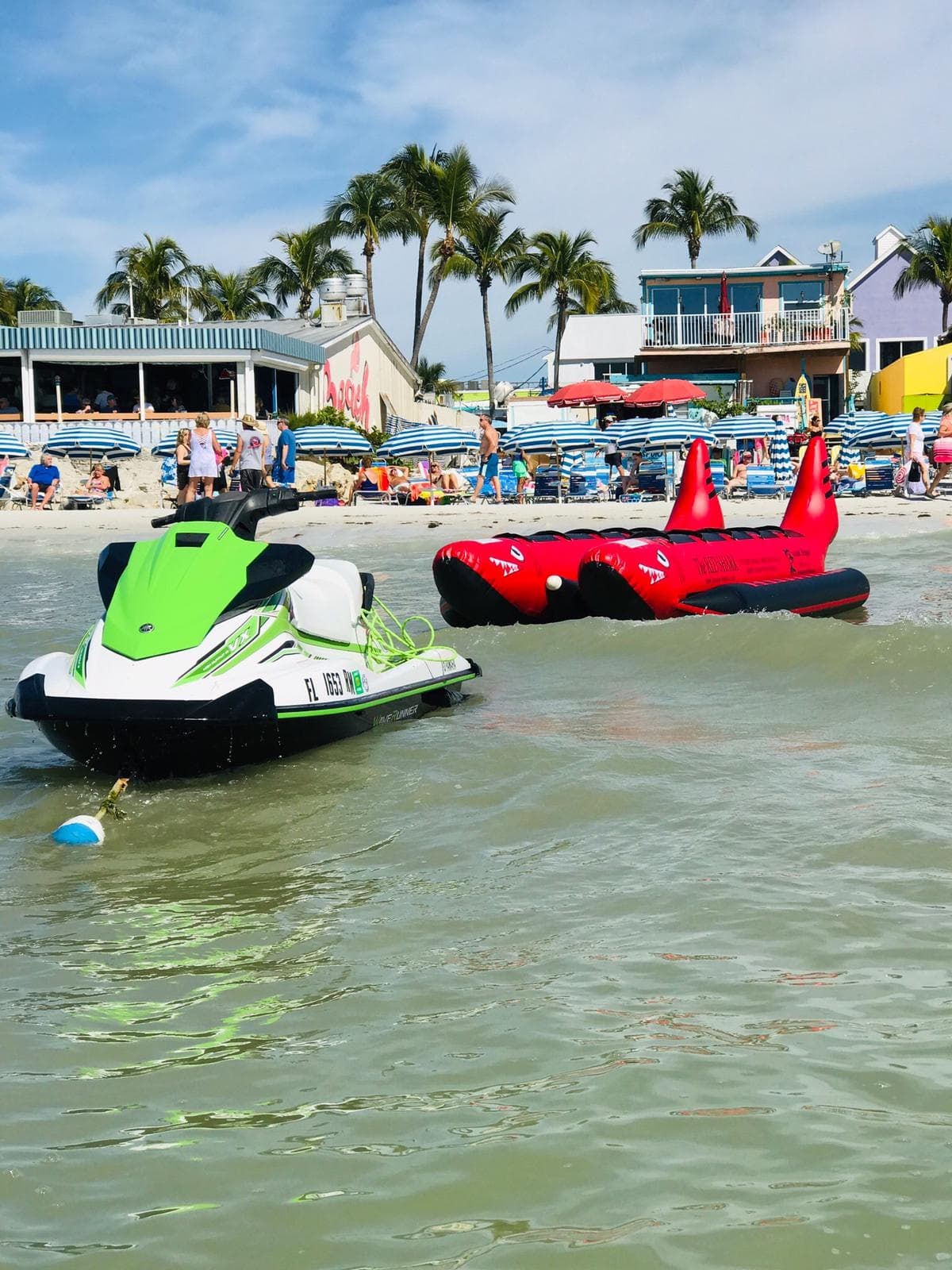 Parked jet skis near crowded sandy beach with palm trees, rental setup