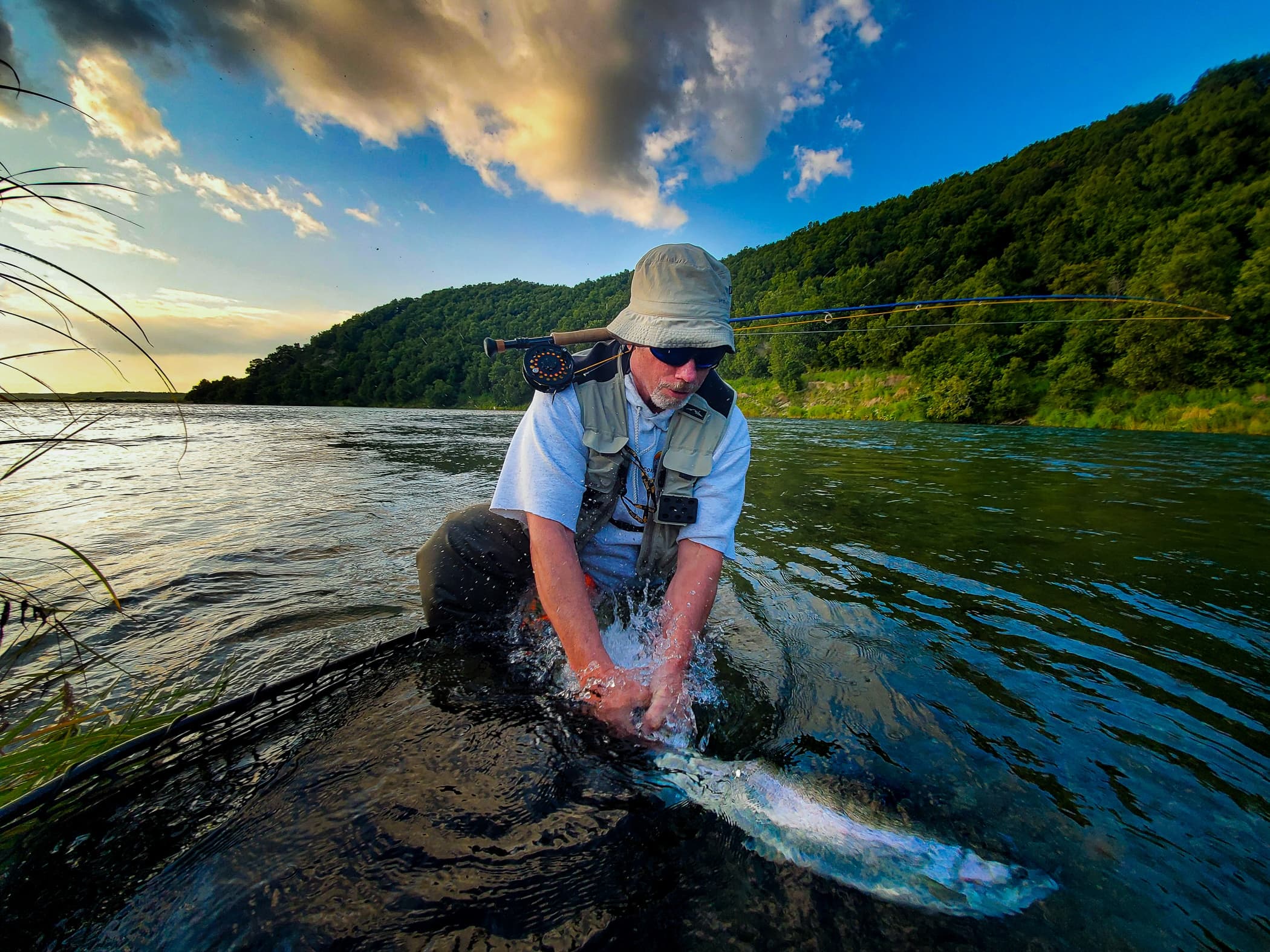Fly angler releasing steelhead in river under dramatic sky