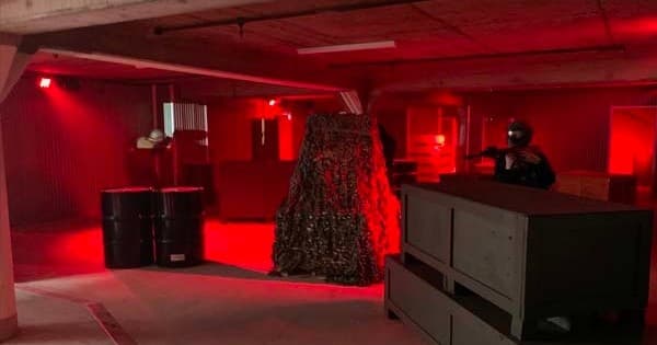 Red-lit indoor airsoft field with player behind barricade in action