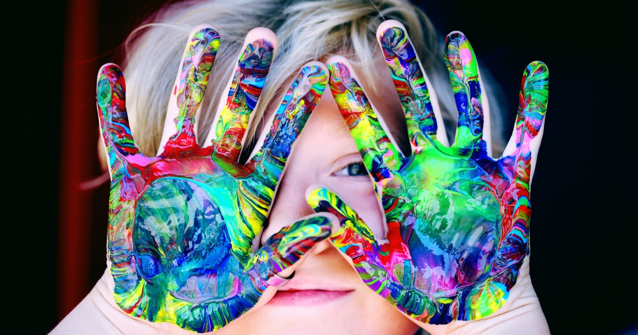 Child showing colorful painted hands covering face, indoor art activity