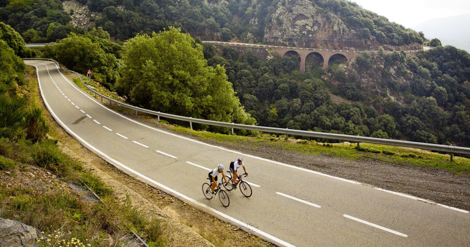 Group road cyclists riding winding viaduct road through hills