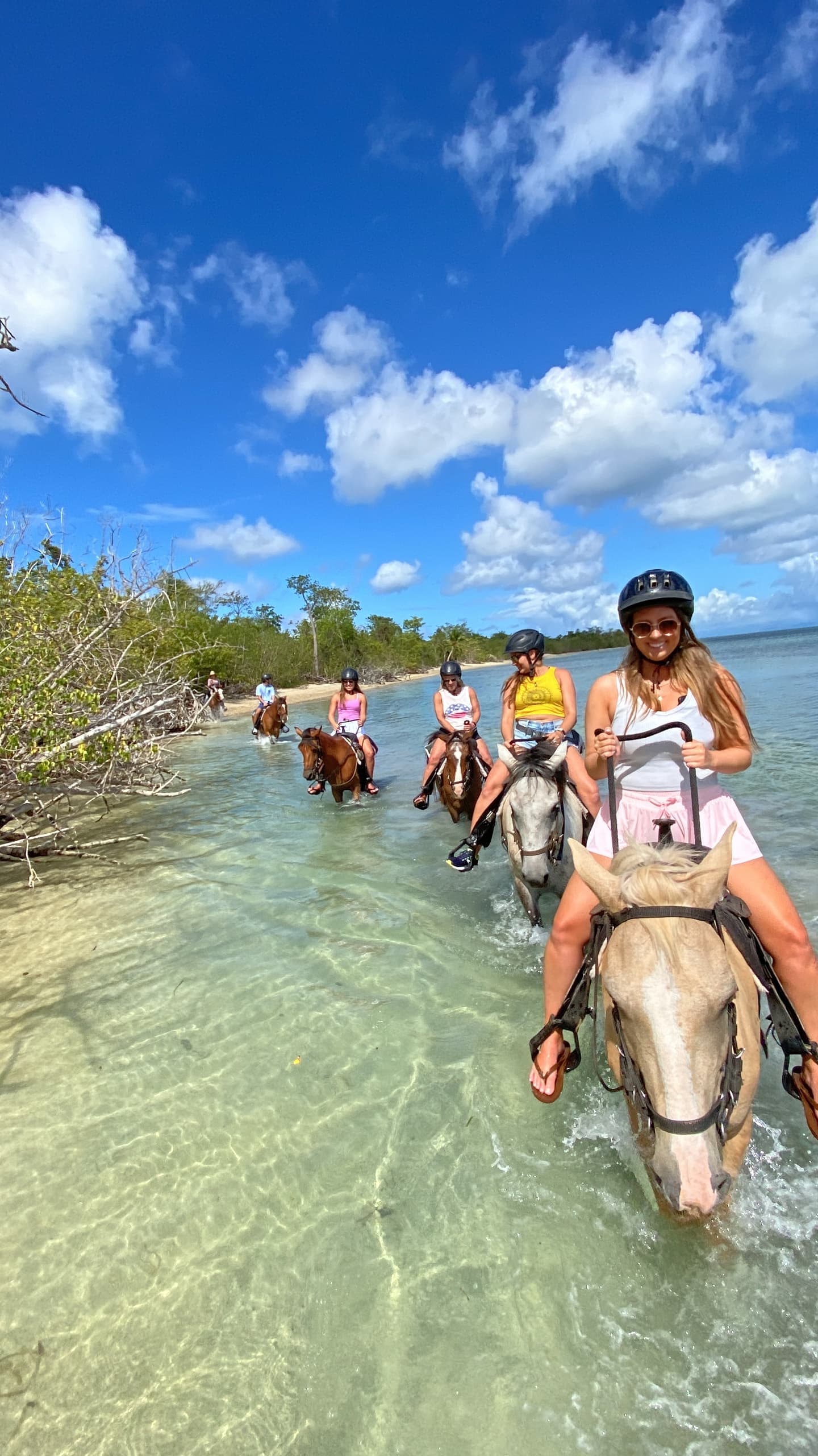 Group horseback riding through shallow tropical water under blue sky