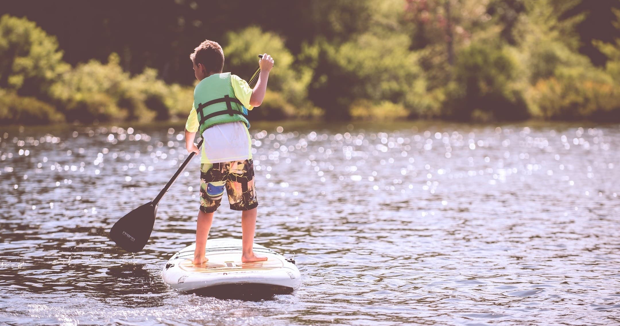 Child paddleboarding on sunny lake shore with life vest