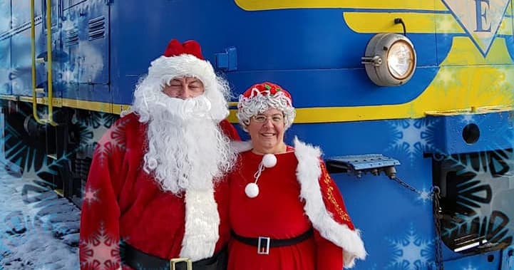 Santa and Mrs. Claus posing beside blue scenic train at depot