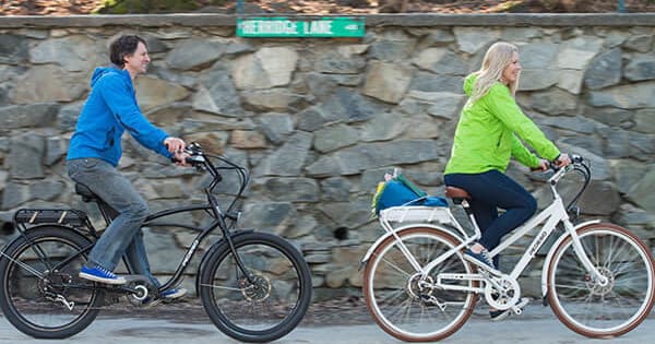 Two cyclists riding e-bikes along stone wall in suburban trail