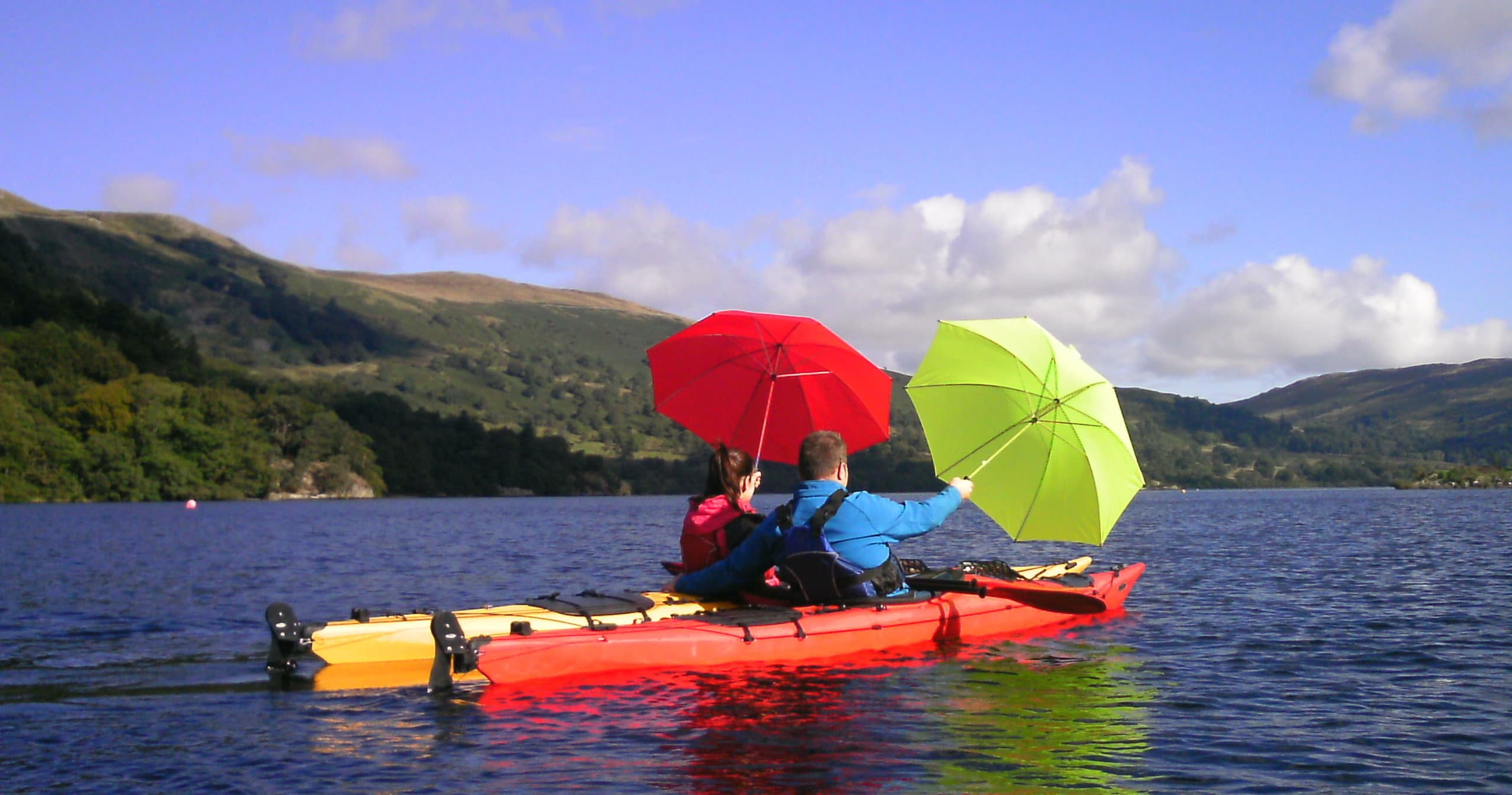 Two kayakers with colorful umbrellas paddling on scenic mountain lake