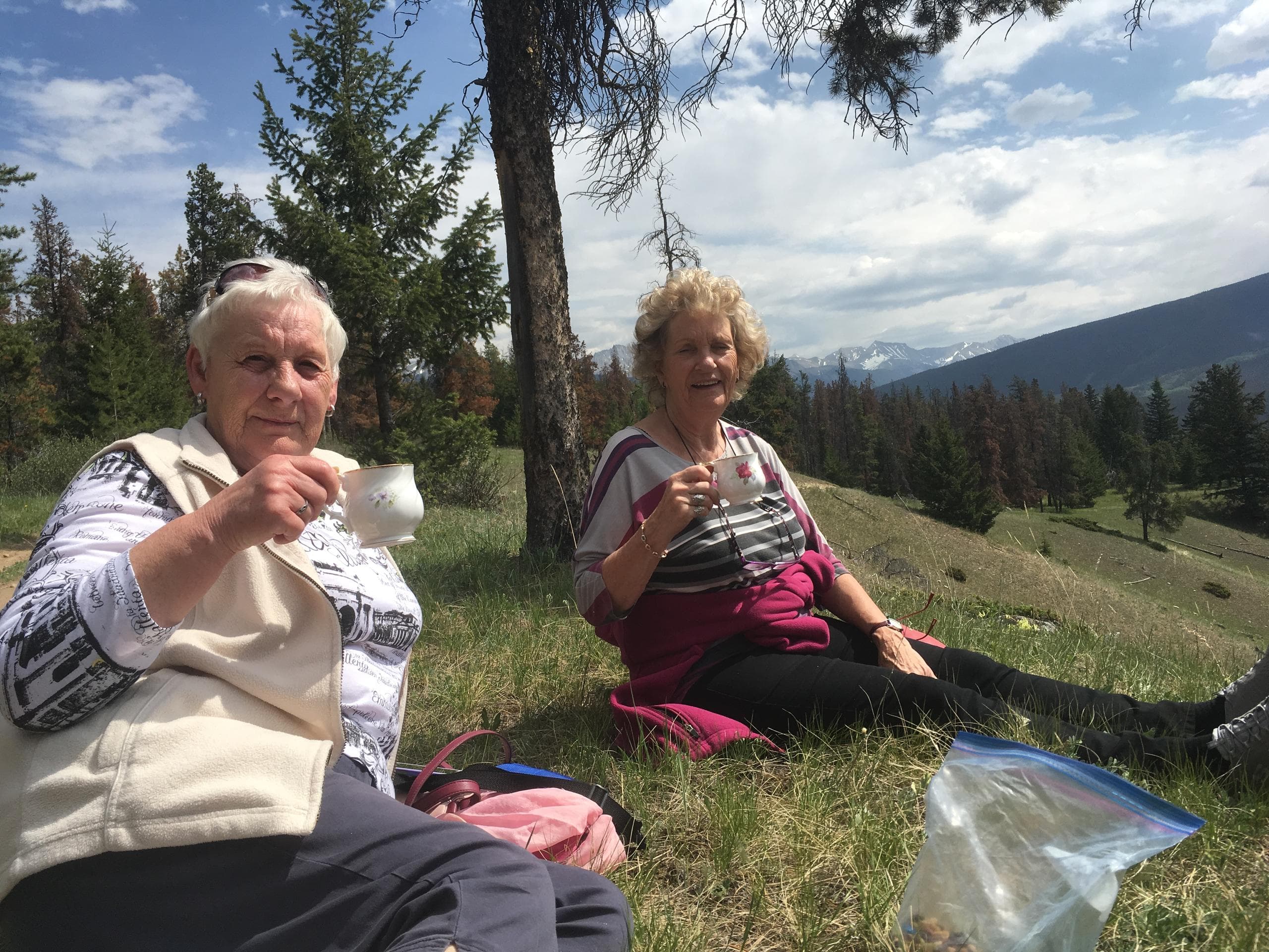 Two women picnic on grassy alpine meadow with mountain views