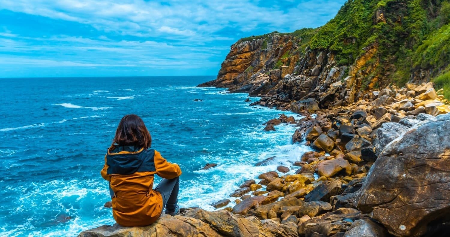 Hiker sitting on rocky cliff overlooking turquoise sea, coastal hike