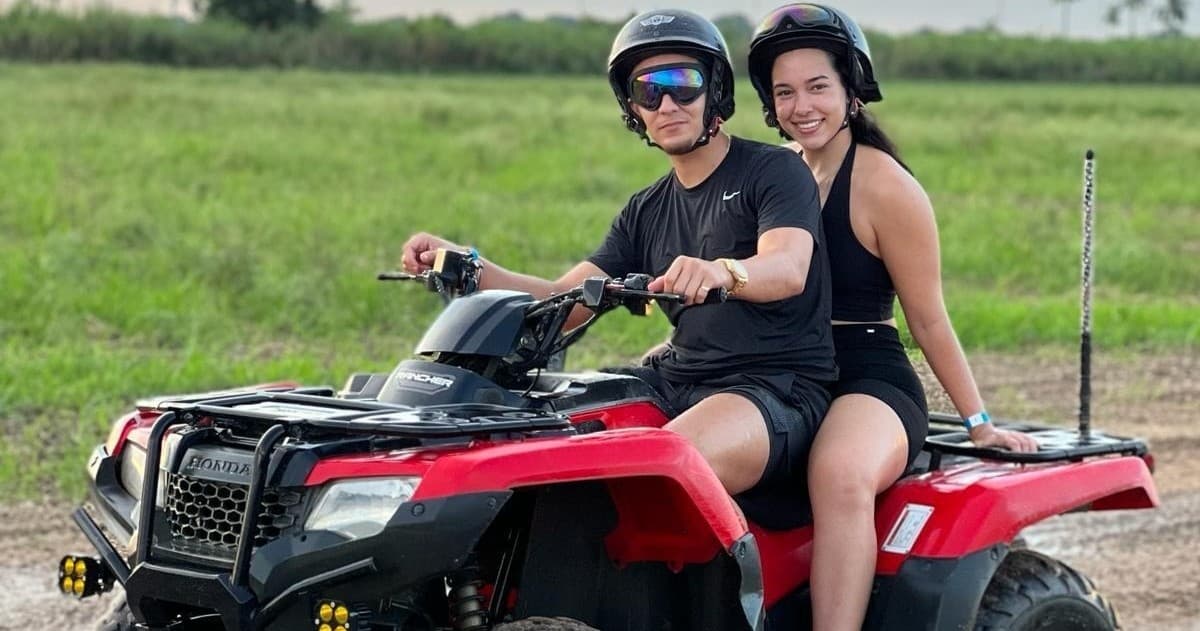 Two riders on a red ATV in grassy tropical field, helmets