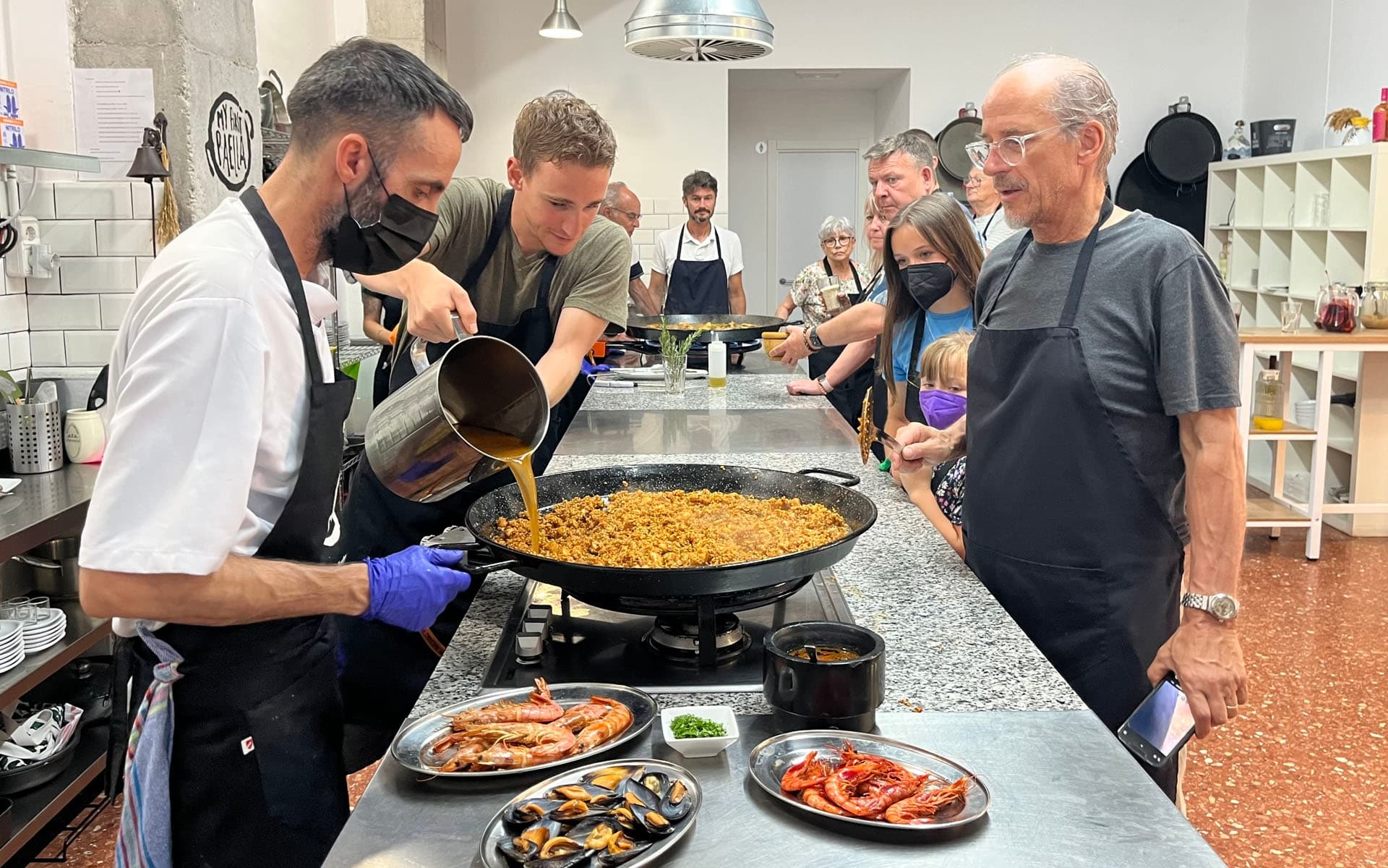 Chef pouring broth into large paella pan during cooking class