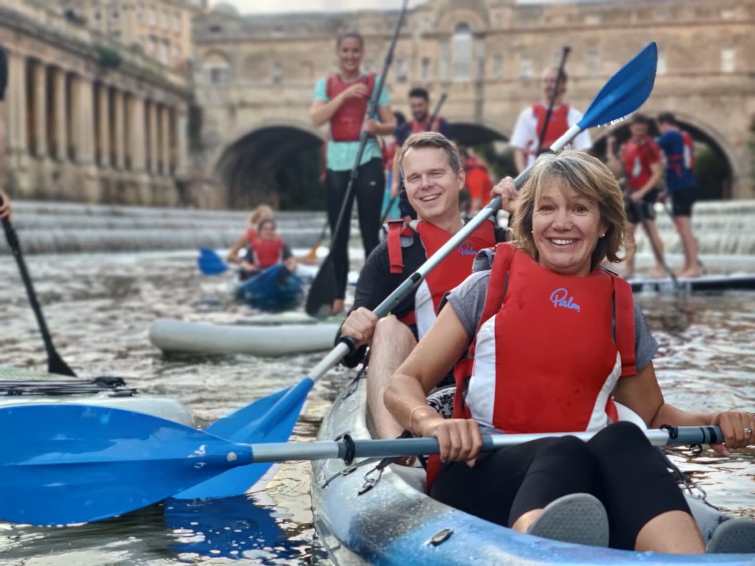 Two kayakers smiling with paddles near historic river bridge