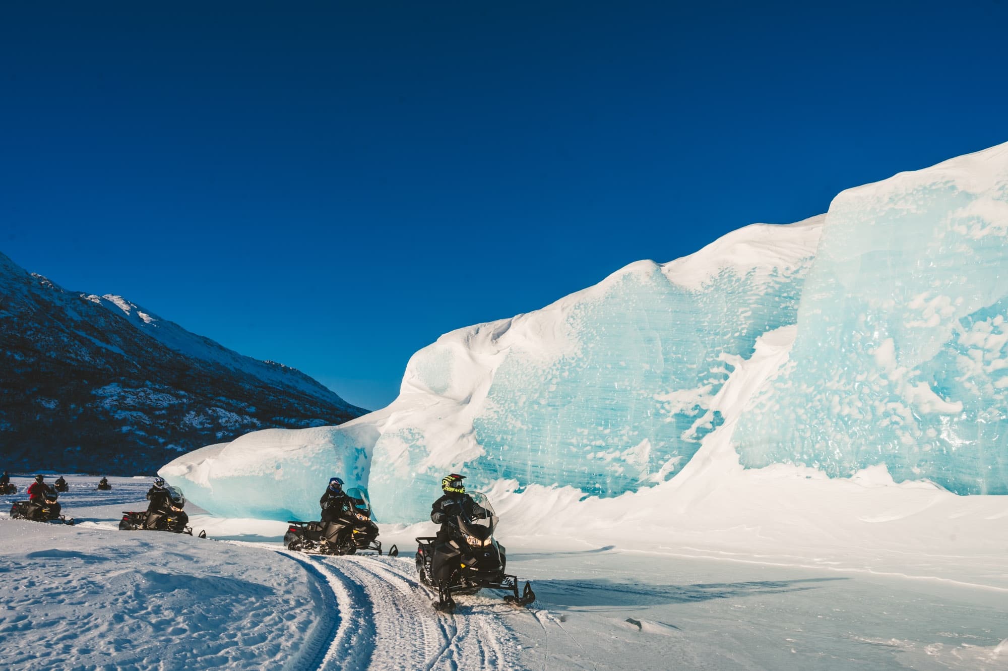 Snowmobile convoy beside towering blue ice formations