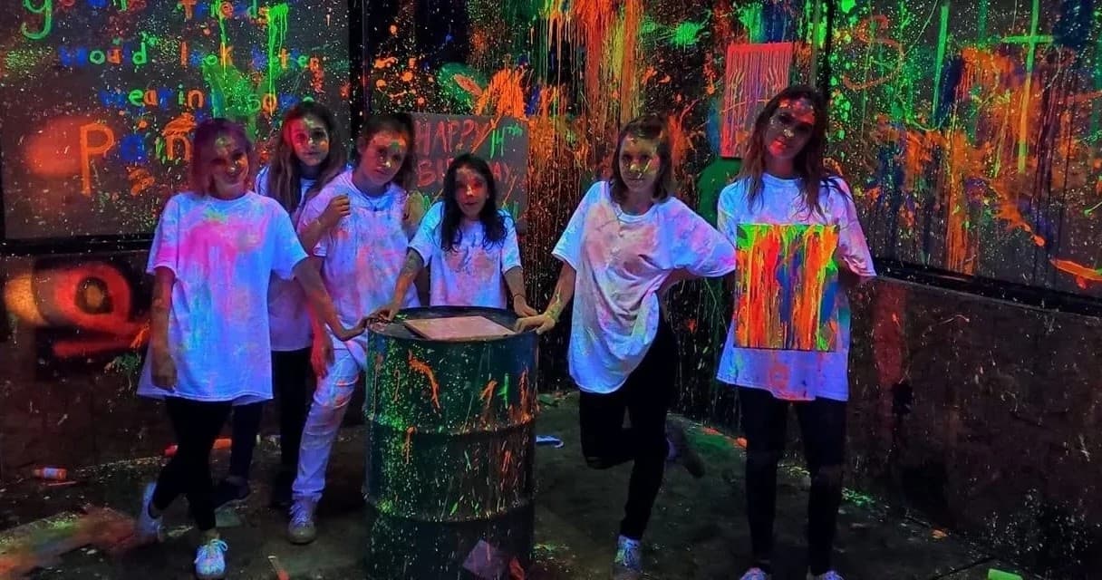 Group in indoor glow paint rage room posing with painted barrel