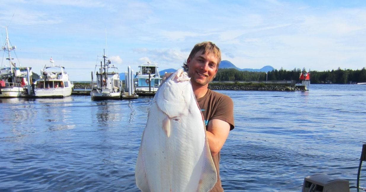 Fisherman holding large halibut in Alaska harbor with boats
