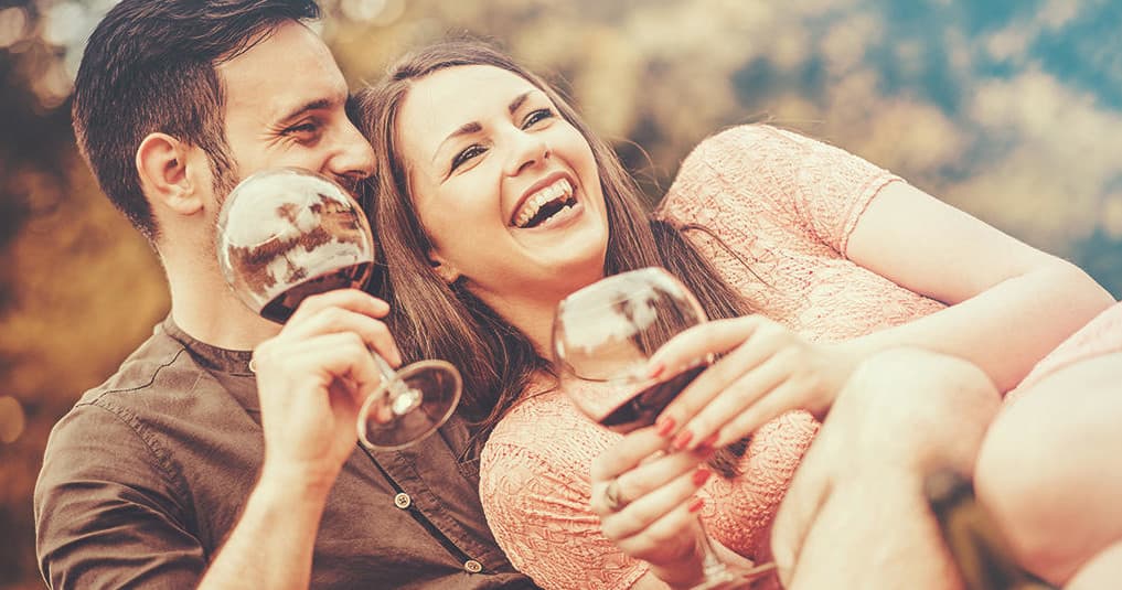 Couple laughing while sipping wine outdoors on picnic grass