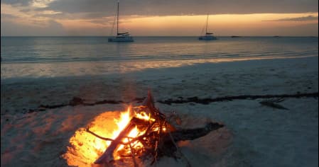 Beach bonfire at dusk with anchored sailboats and shoreline
