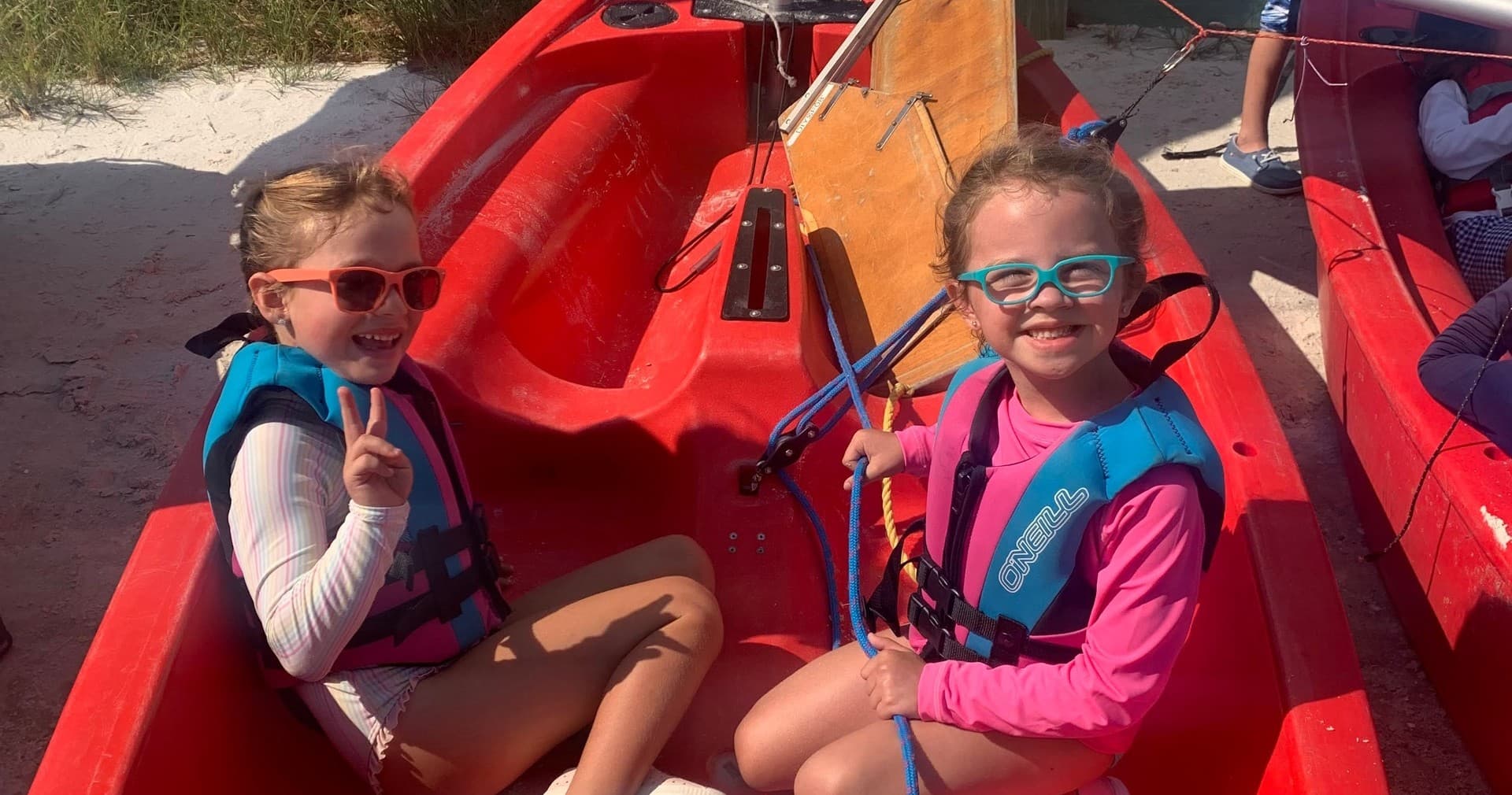 Kids seated in red kayaks on sandy beach smiling with lifejackets