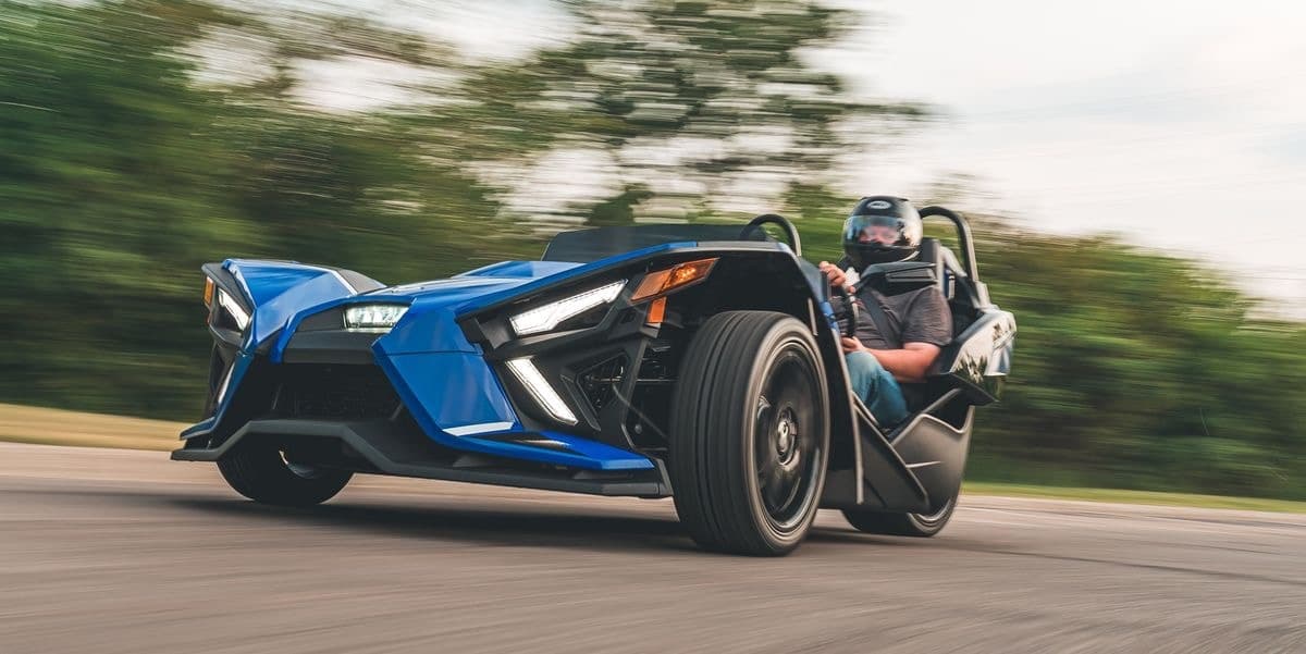 Blue Polaris Slingshot cruising a tree-lined country road