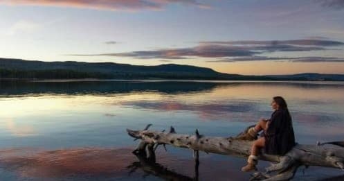 Solo traveler seated on log by calm lake at sunset, peaceful vista