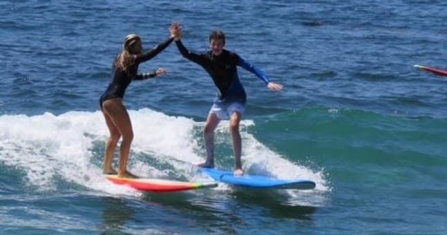 Two surfers balancing on longboards, high-fiving on small ocean waves
