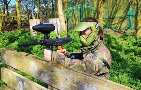Paintball player in camo aiming marker behind wooden barricade