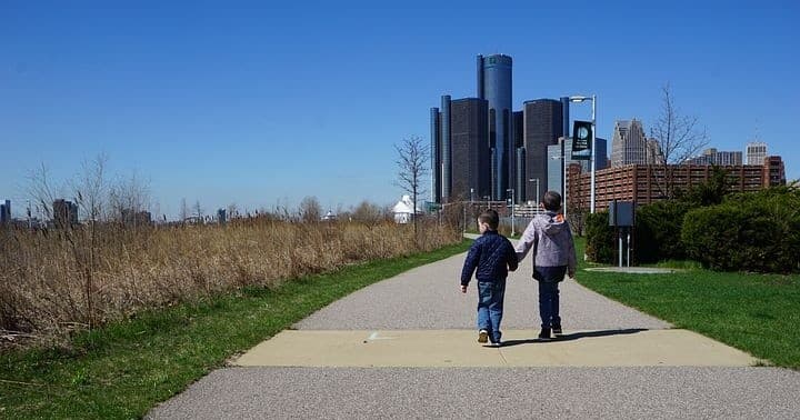 Two people walking on riverside path toward Detroit skyline urban park