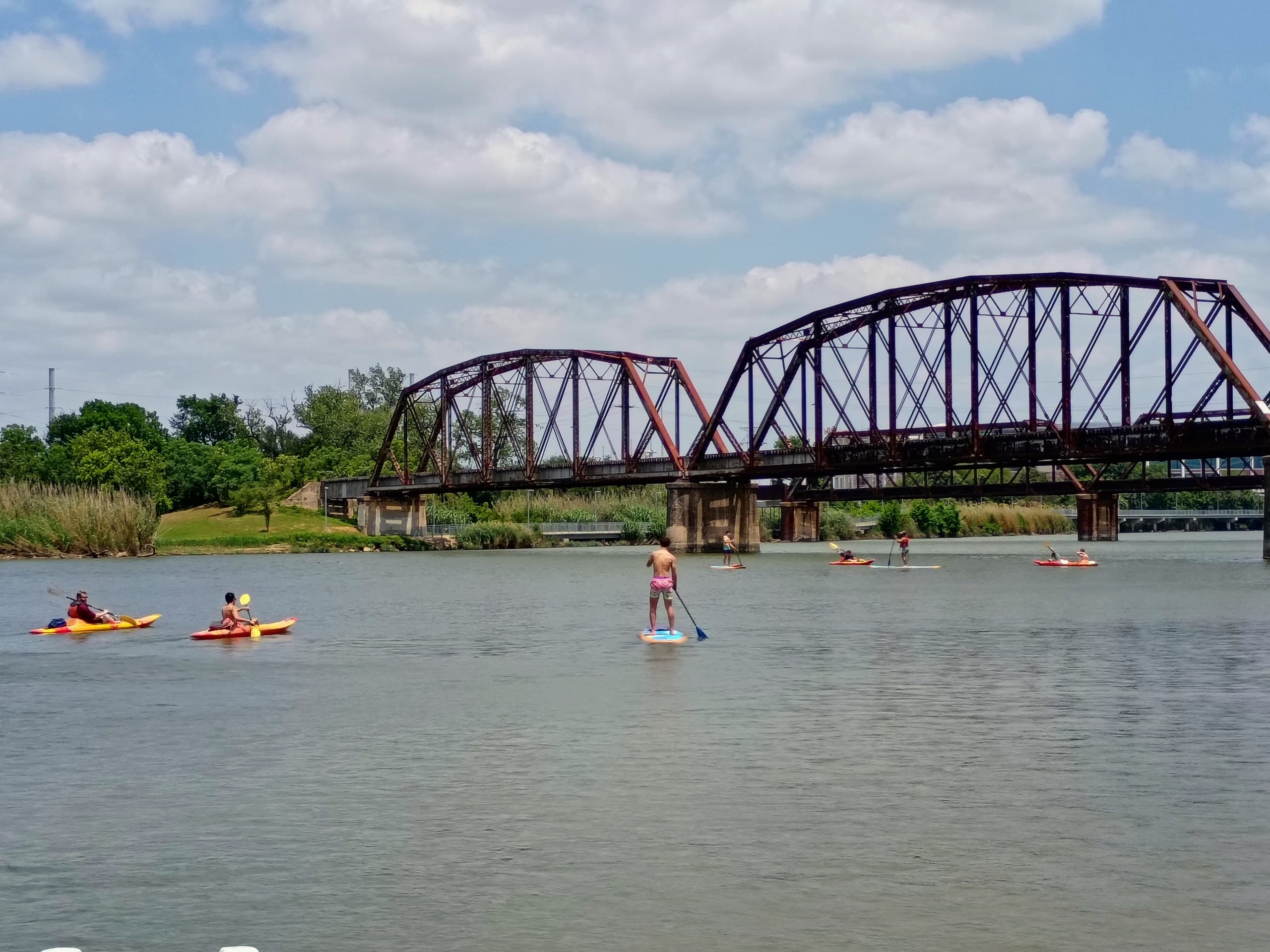 Paddleboarders and kayakers near an iron bridge on calm river